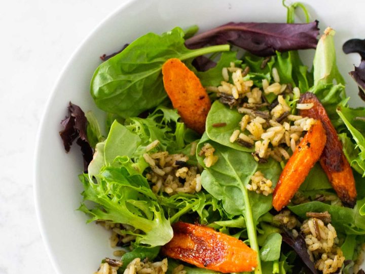 A serving of crispy rice salad shows green lettuce, roasted carrots, fried wild rice in a white bowl.
