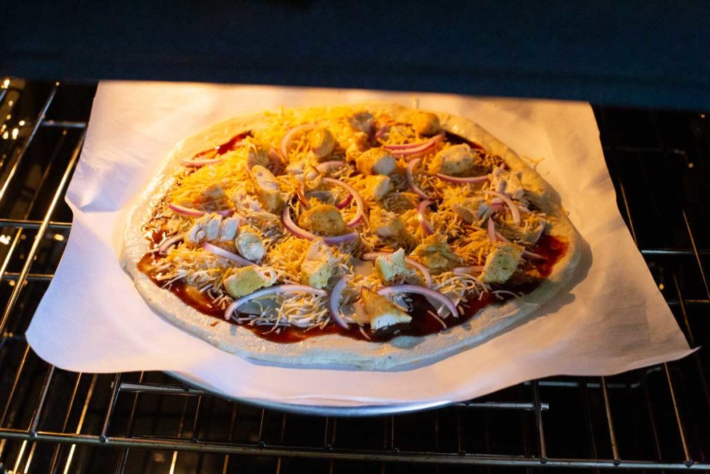 A barbecue chicken pizza is baking in the oven on parchment paper and a metal pizza pan.