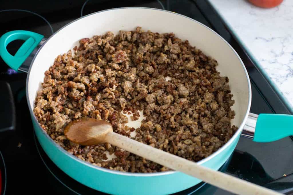 Step 1: Brown the sausage in a skillet. A wooden spoon is shown crumbling the sausage as it cooks.