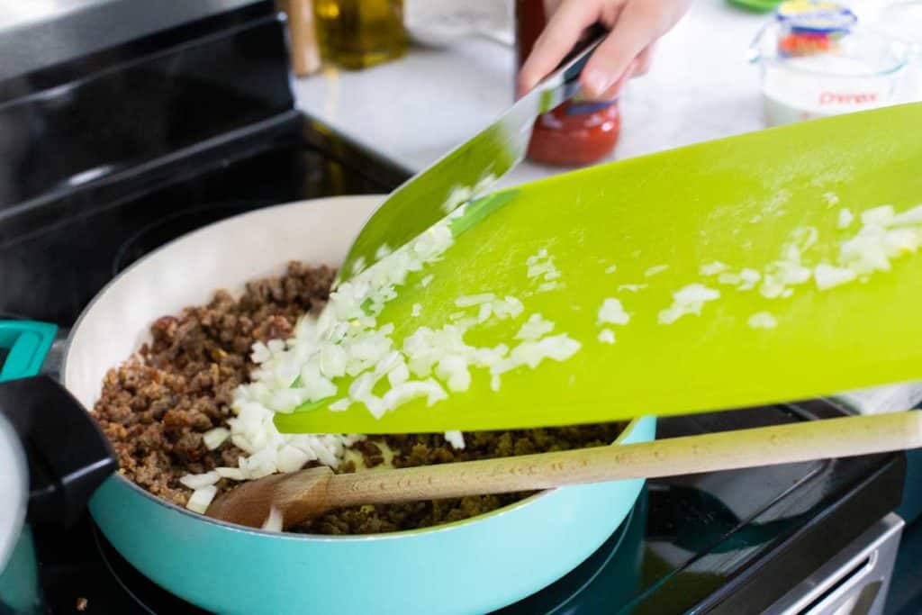 Step 2: Add the chopped onions and garlic. A hand is scraping onions from a cutting board into the skillet.