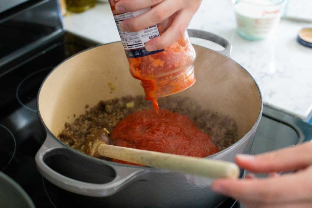 Step 3: Add the jarred tomato sauce to the skillet. A hand is holding the glass jar.