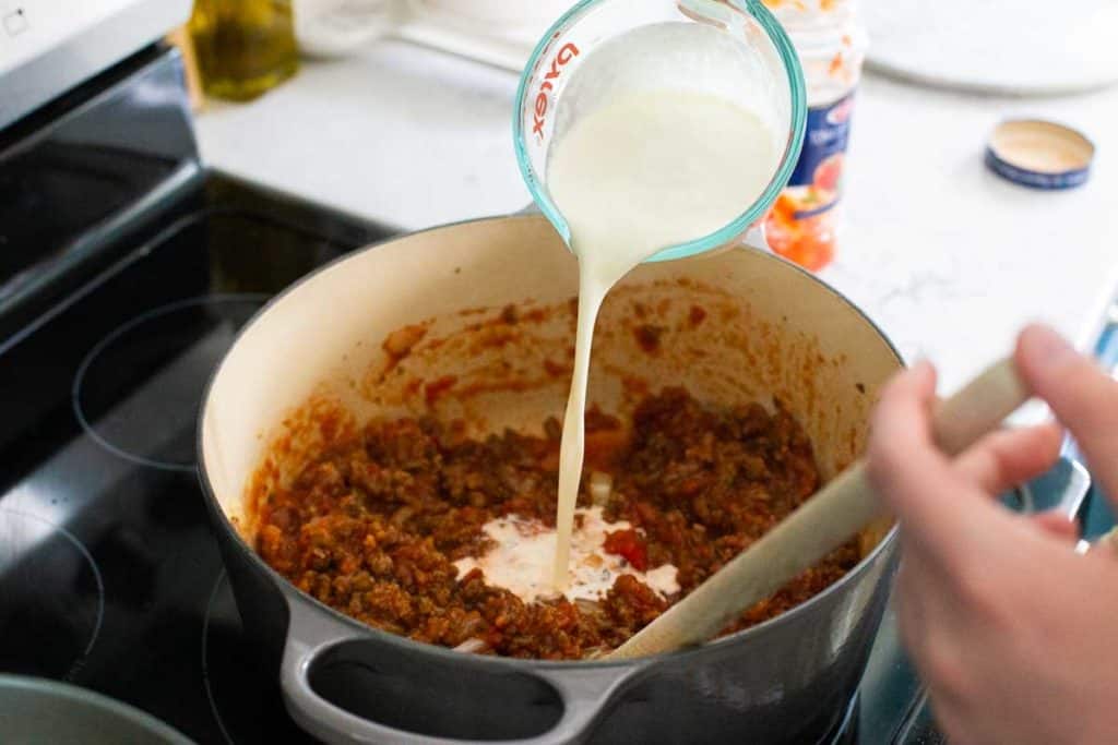 Step 4: Add heavy cream to the sauce. A measuring cup is seen drizzling the cream into the tomato sauce.