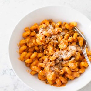 A white bowl holds curly pasta coated in a creamy tomato sauce with crumbled sausage and parmesan sprinkled on top.