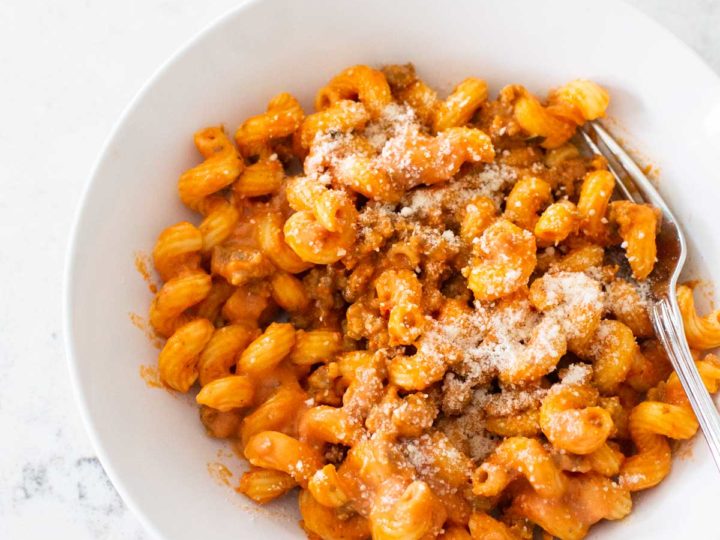 A white bowl holds curly pasta coated in a creamy tomato sauce with crumbled sausage and parmesan sprinkled on top.