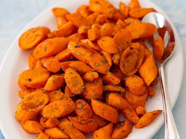 The oven roasted carrots have golden brown edges and are being served on a white platter with a serving spoon.