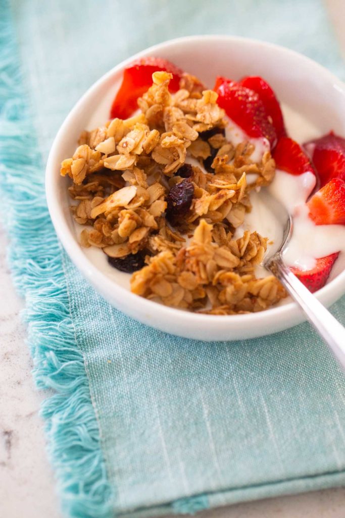 The baked granola is in a bowl with yogurt and fresh strawberries and sits on a blue napkin.