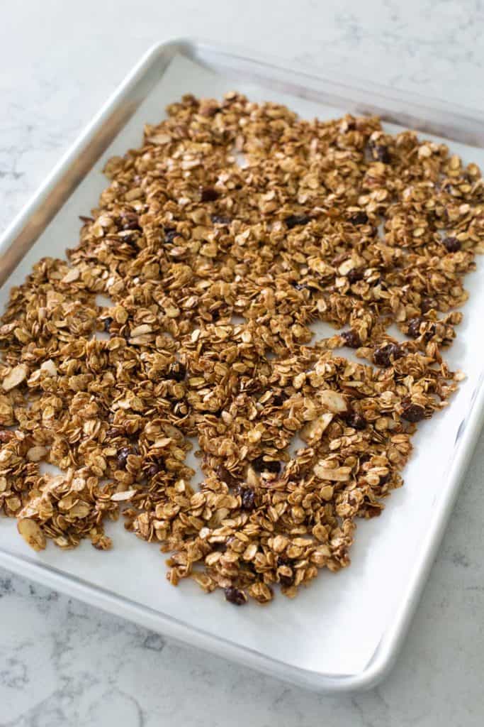 The baking sheet is lined with parchment and has the granola spread in an even layer.