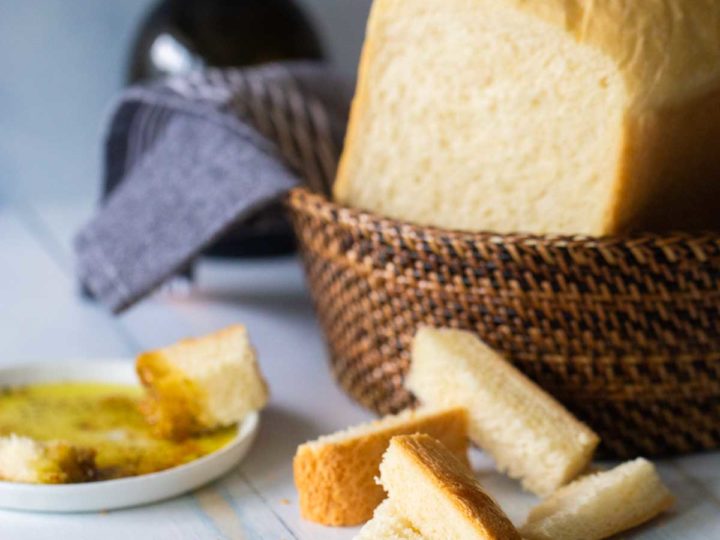 A loaf of bread machine Italian bread sits in a basket next to a few sample wedges near a plate of olive oil for dipping.