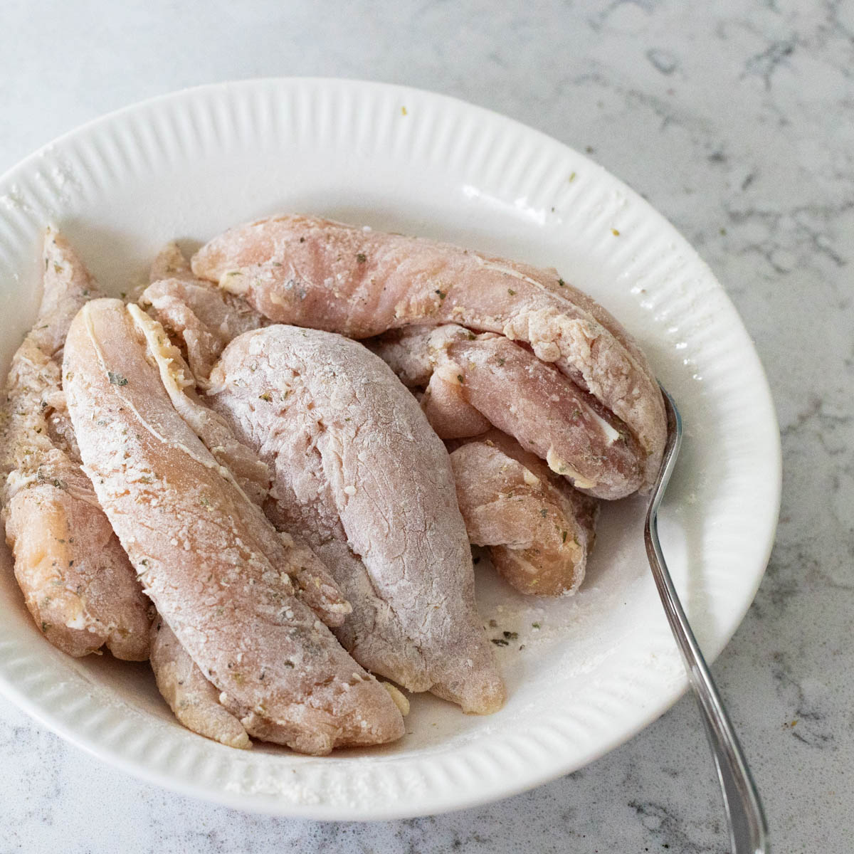 The chicken tenderloins have been seasoned and tossed in flour in a small bowl.