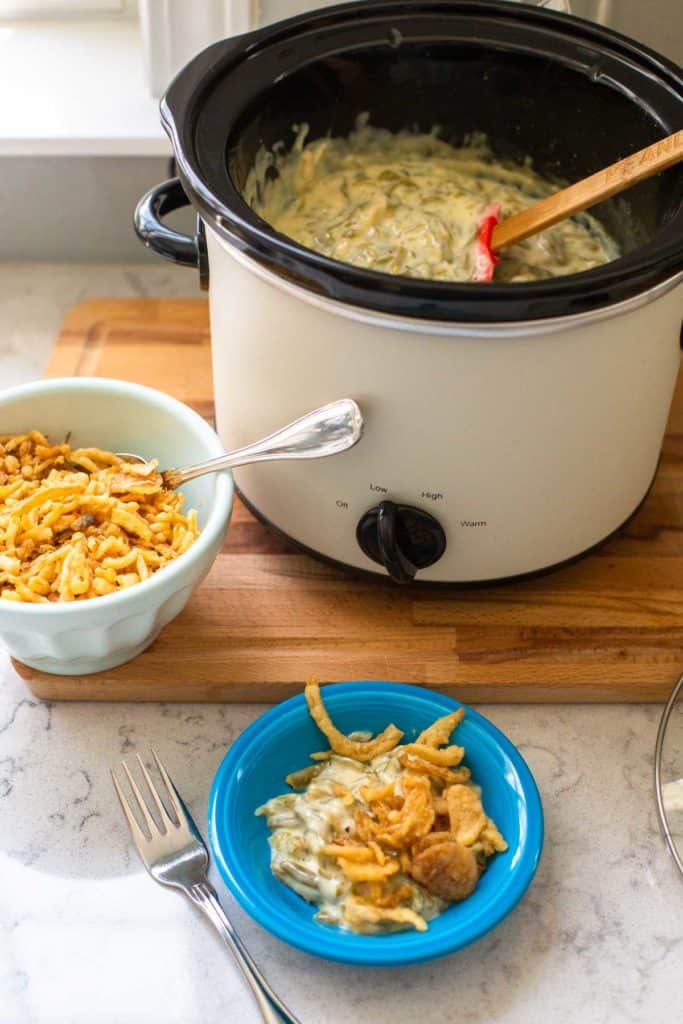 The open crockpot has a serving spoon ready and sits next to a portion of green bean casserole.