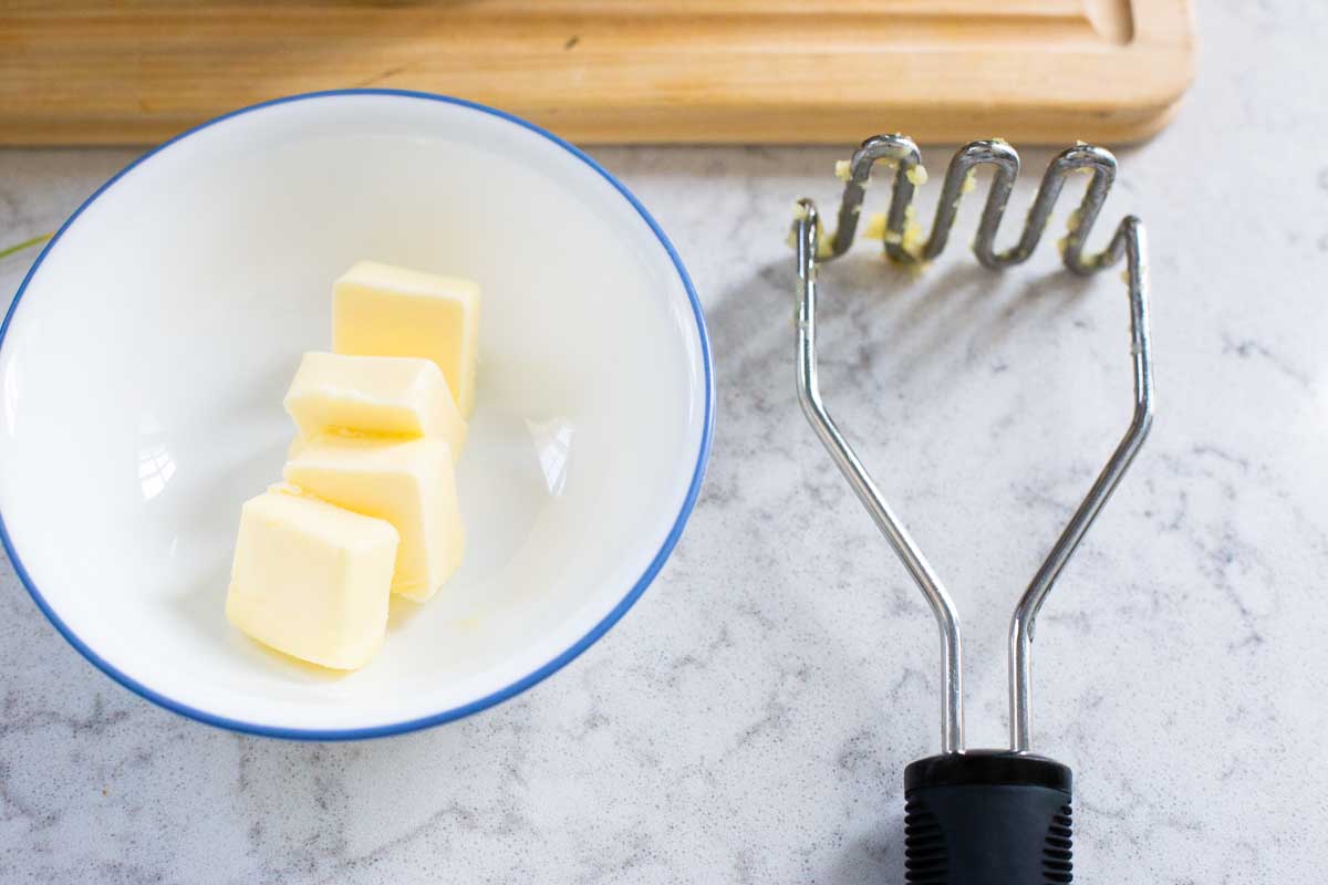 Softened butter has been sliced into pieces and waits in a small bowl next to a potato masher.