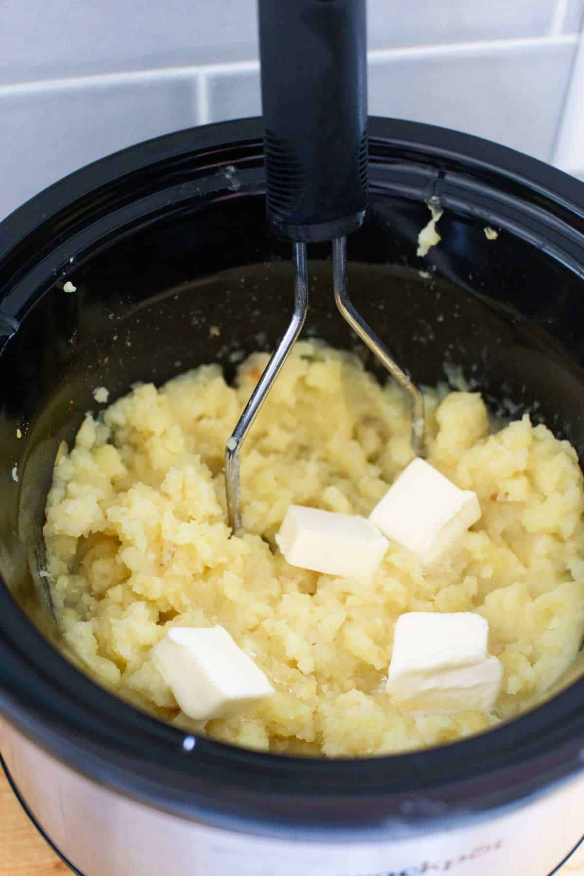 The cooked potatoes are being mashed by a masher and cubes of butter are melting into the potatoes.