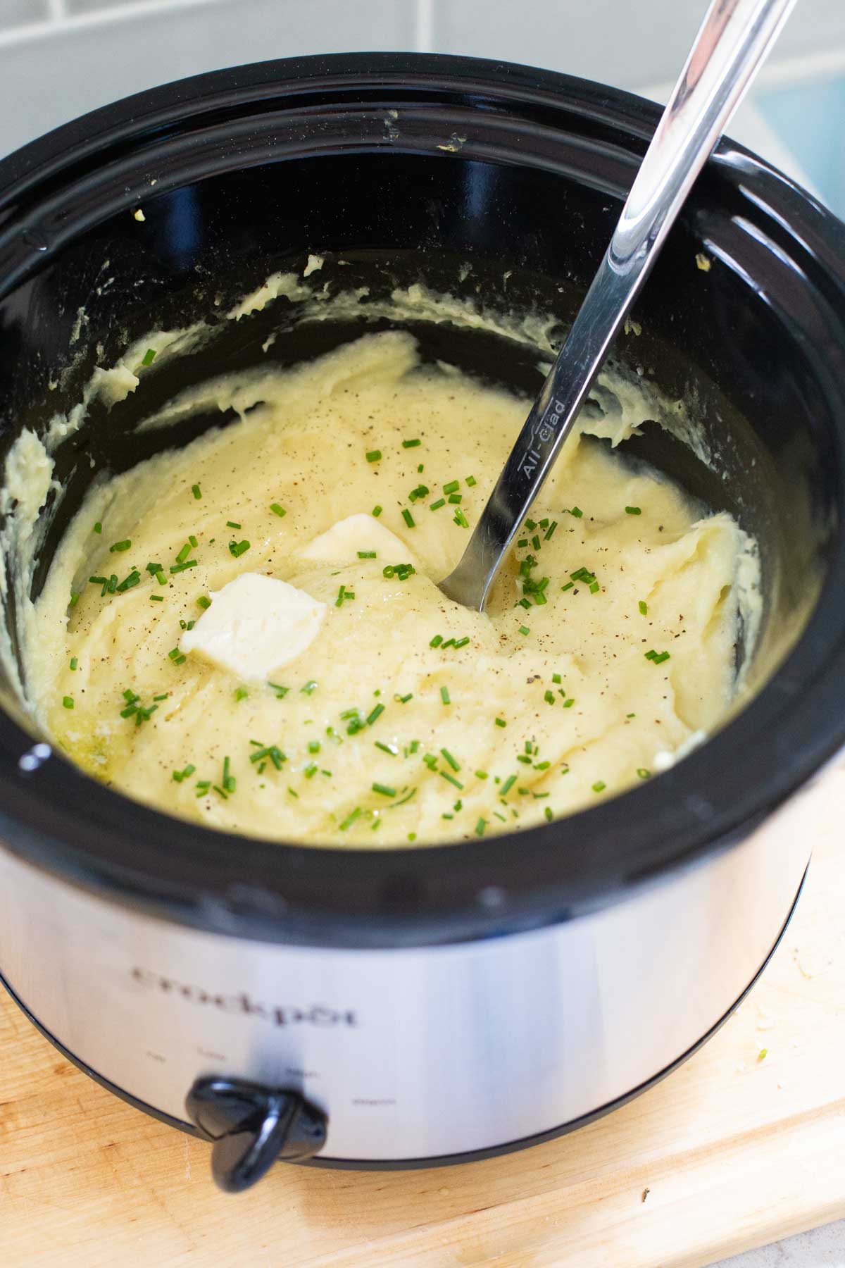The finished mashed potatoes are in the Crockpot with a serving spoon, a pat of butter, and fresh chopped chives.