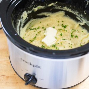 A silver Crockpot has finished mashed potatoes that are sprinkled with fresh chives and two pats of butter.