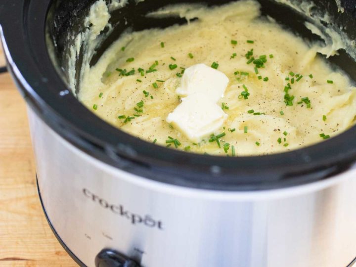 A silver Crockpot has finished mashed potatoes that are sprinkled with fresh chives and two pats of butter.