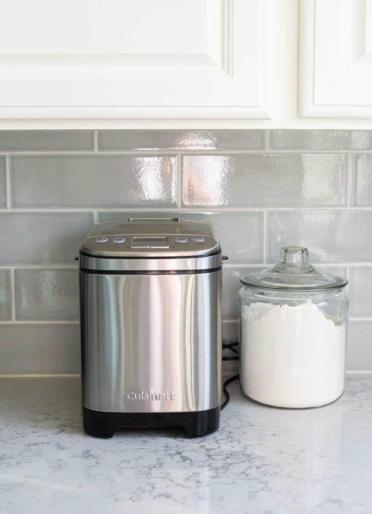 The compact Cuisinart bread machine is on a kitchen counter underneath a kitchen cabinet.