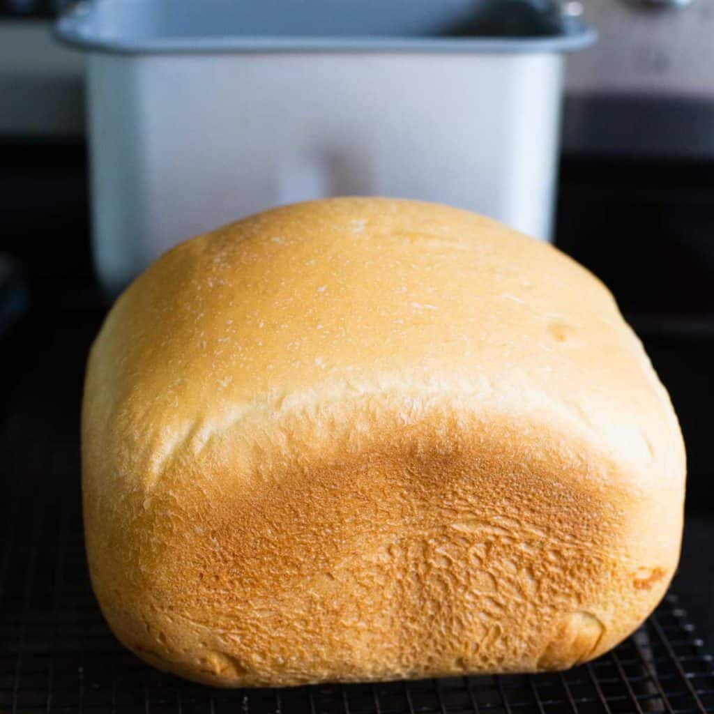 A loaf of bread machine bread cools on a wire rack with a bread pan in the background.