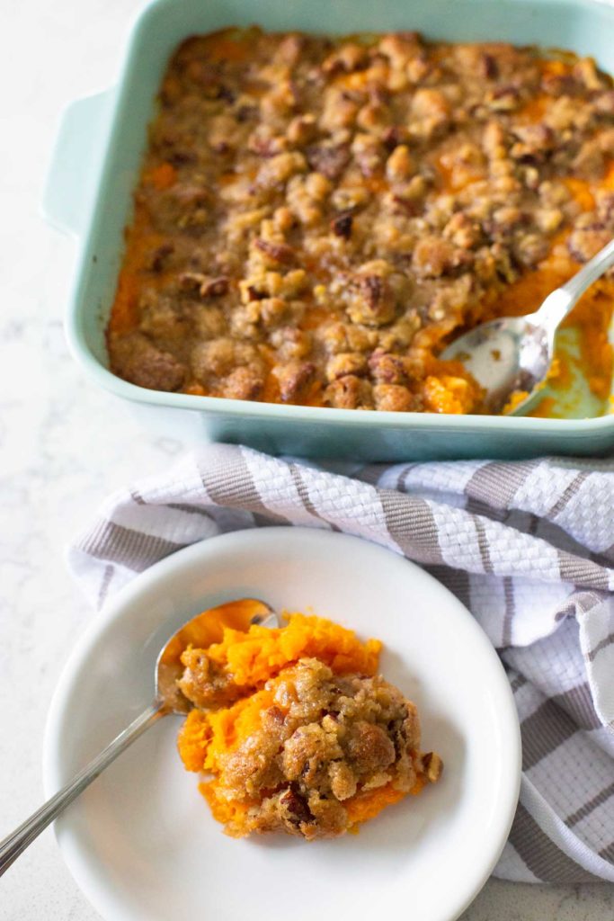 A serving of sweet potato casserole sits on a plate with a spoon in front of the baking dish.