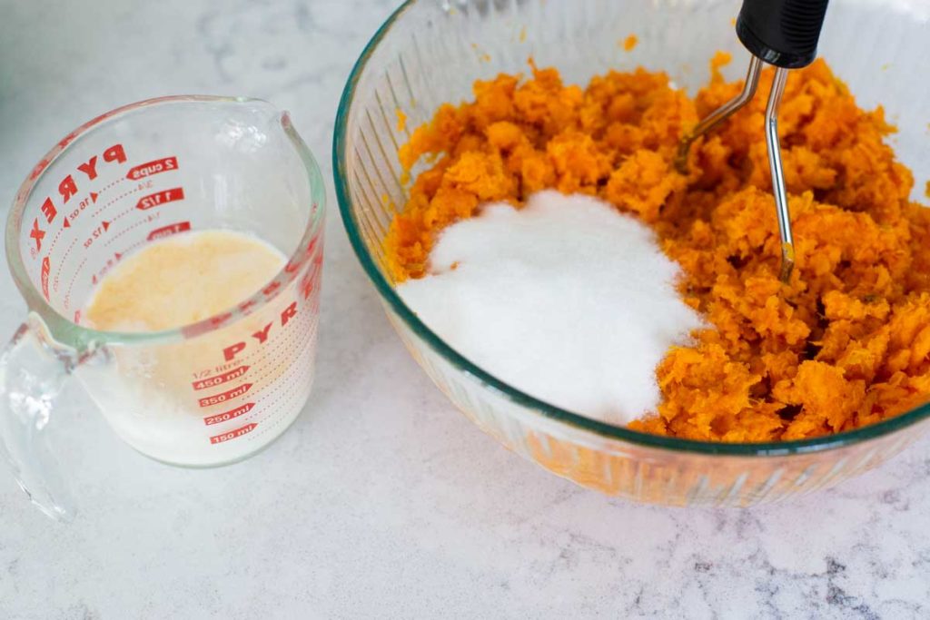 Sugar and half and half in a measuring cup are getting mixed into the mashed sweet potatoes in a mixing bowl.