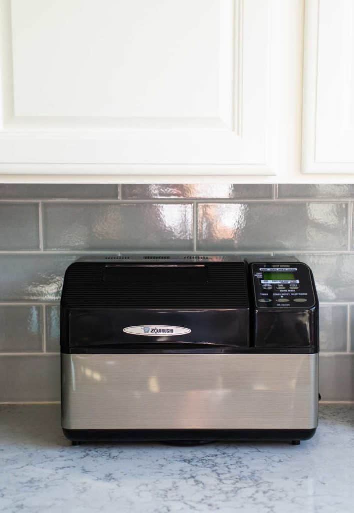 The Zojirushi bread machine sits on a kitchen counter underneath a kitchen cabinet.