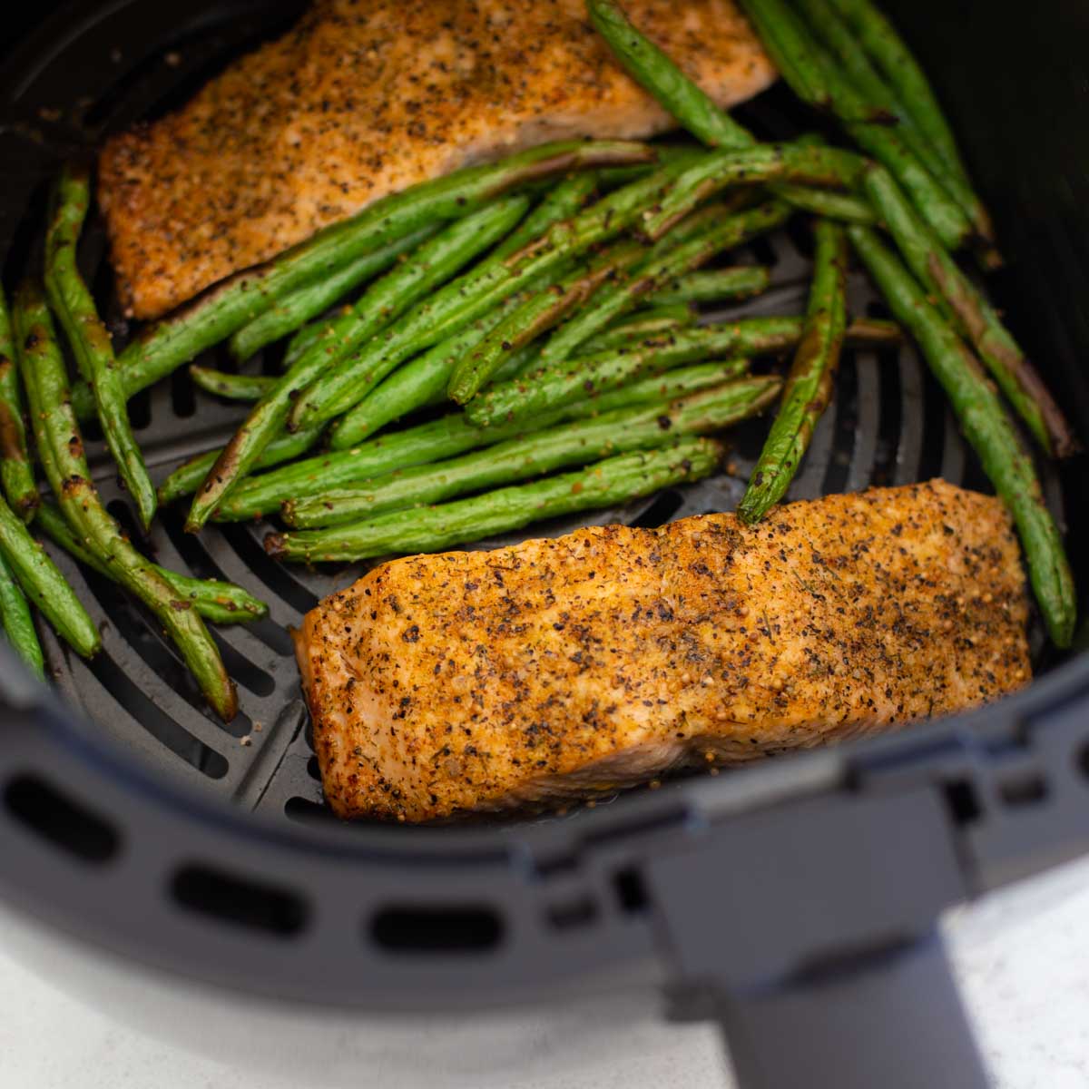 Two salmon filets are surrounded by fresh green beans in an air fryer basket.