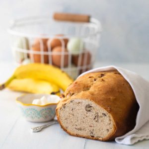 Banana bread wrapped in a tea towel sits next to fresh bananas and a bowl of cream cheese. A basket of eggs in the back.