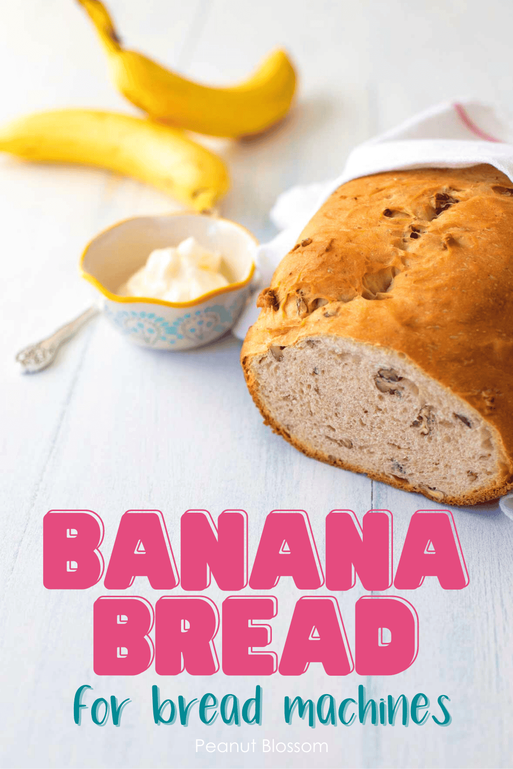 The finished banana bread sits on a table next to a few bananas and a bowl of cream cheese.