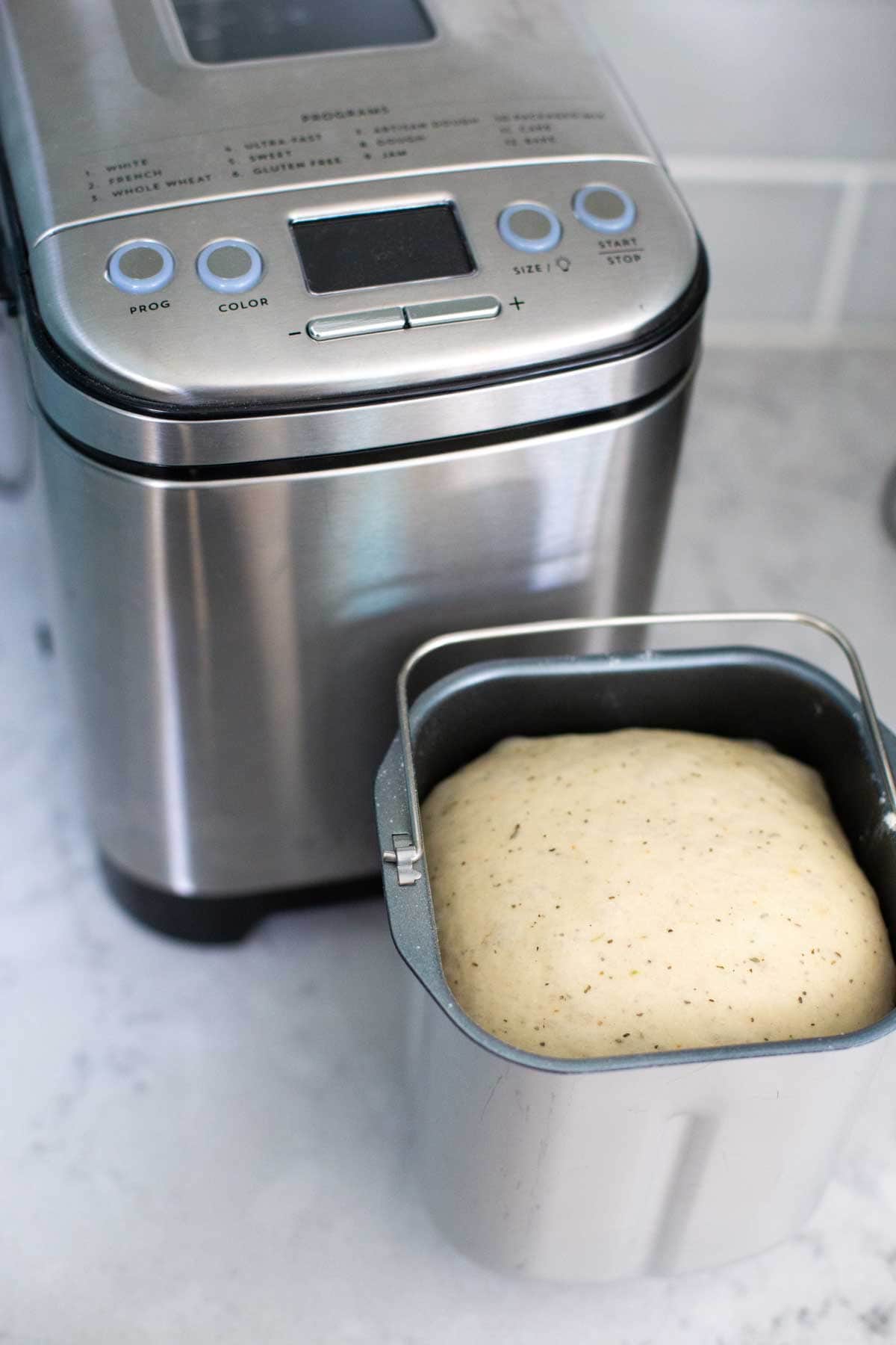 A bread pan filled with focaccia dough sits in front of a Cuisinart compact bread maker.