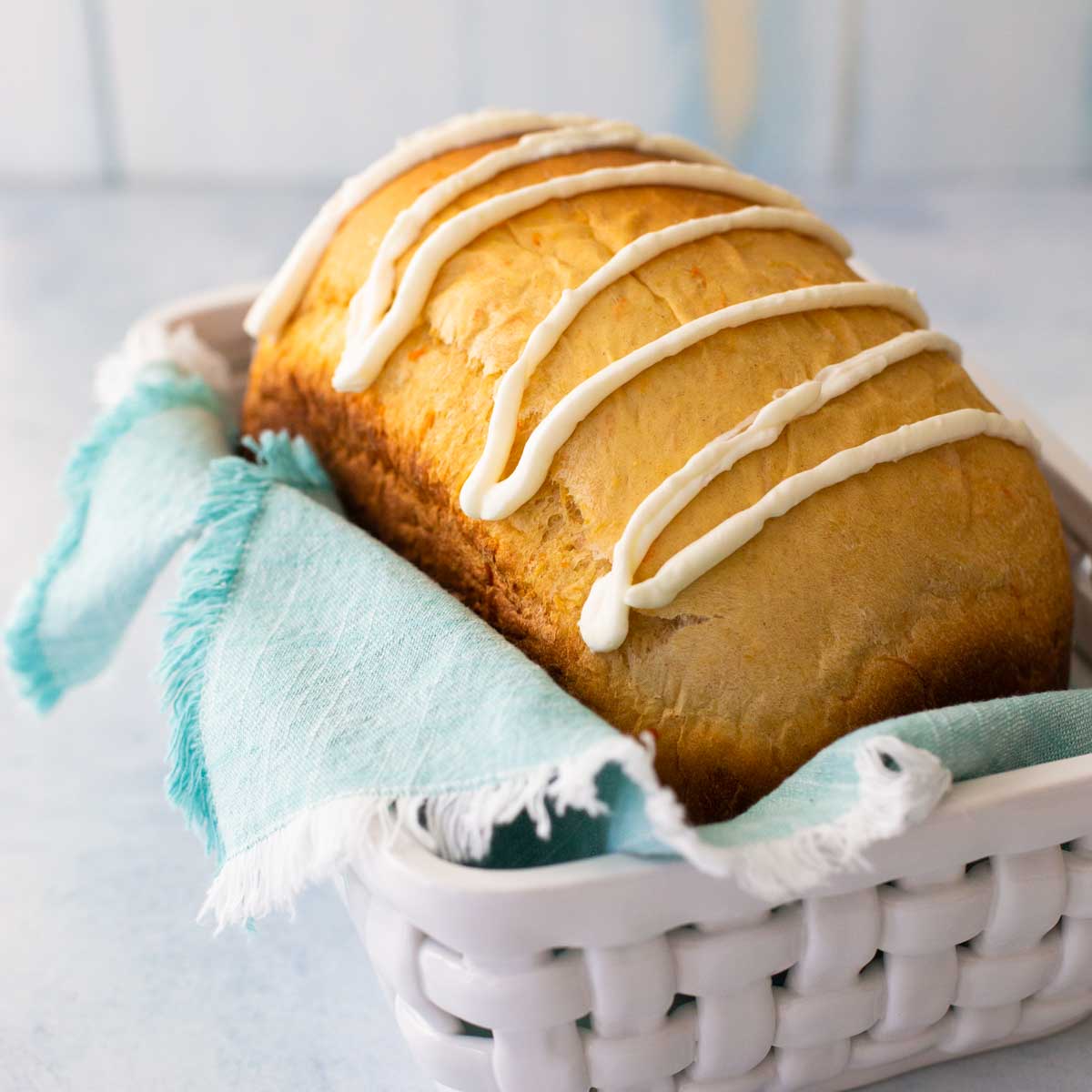 An iced loaf of carrot cake bread sits in a white bread basket with a blue napkin.