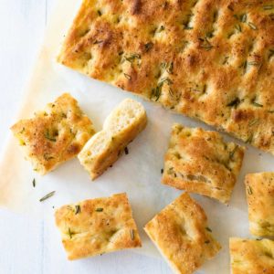 A focaccia bread on parchment paper with several squares cut and scattered.