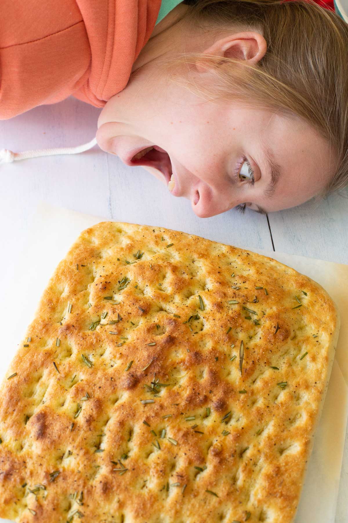 A young girl playfully tries to bite the entire focaccia bread.