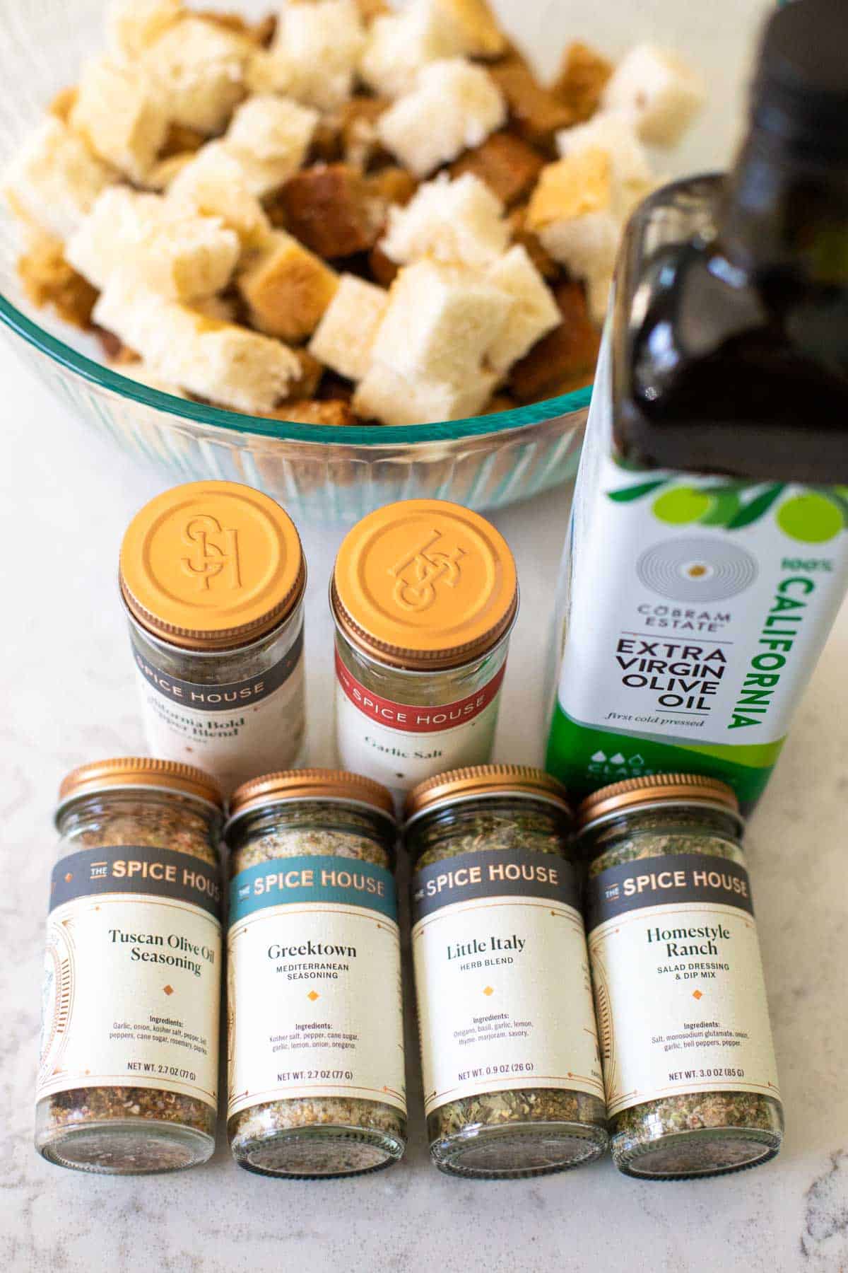 A variety of jars of seasonings that could be used to season the croutons sit next to a jar of olive oil. 