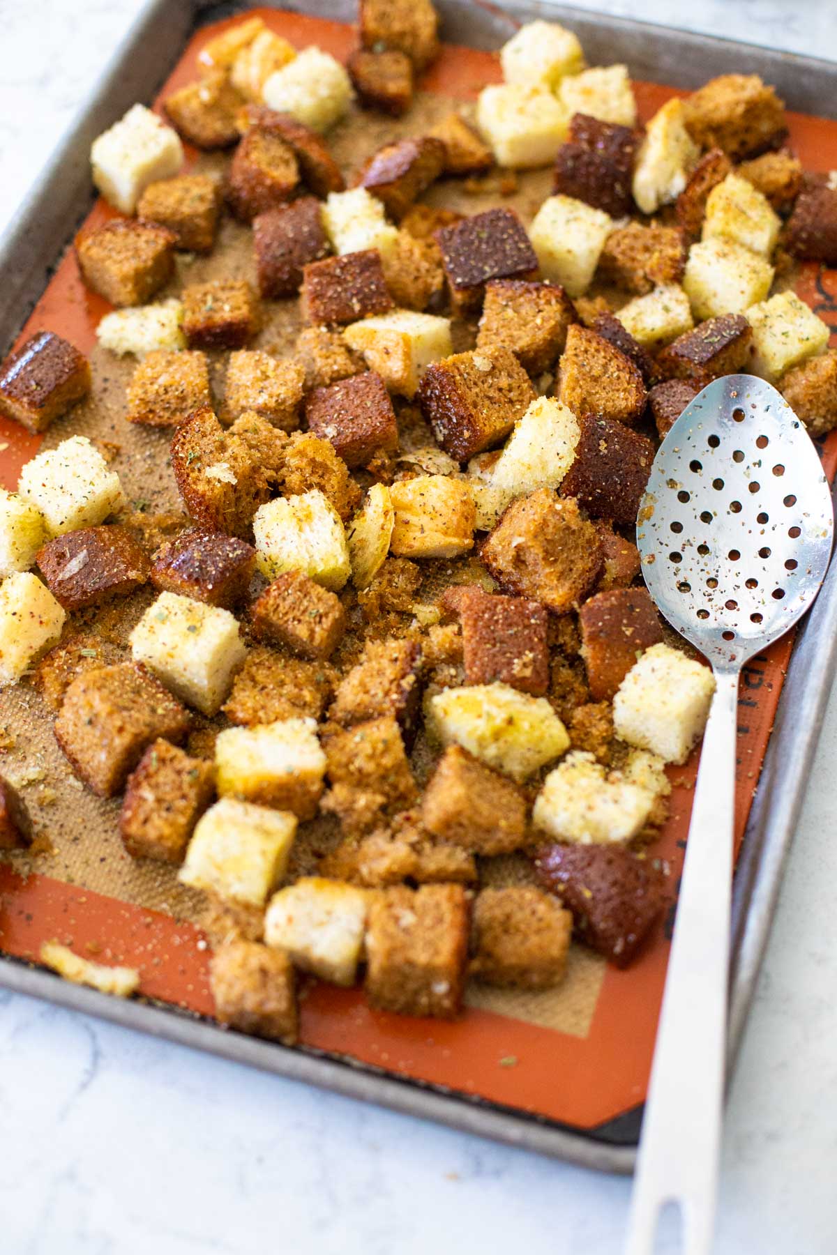The croutons are spread out on a baking sheet next to a metal cooking spoon.