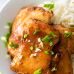 A plate of baked honey garlic chicken thighs on a bed of white jasmine rice sprinkled with green onions.