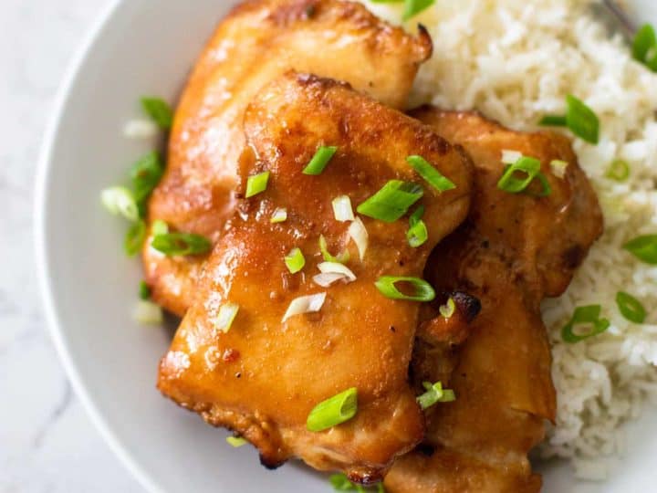A white plate has jasmine rice and a pile of honey garlic chicken thighs sprinkled with fresh green onions.