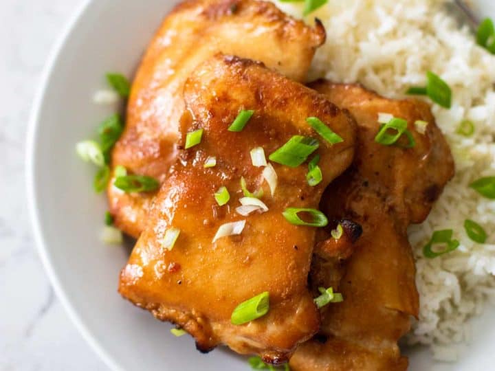 A white plate has jasmine rice and a pile of honey garlic chicken thighs sprinkled with fresh green onions.