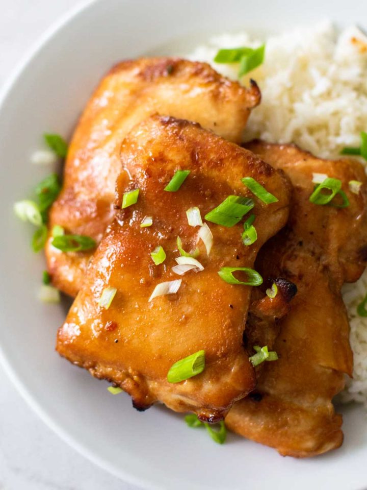 A white plate has jasmine rice and a pile of honey garlic chicken thighs sprinkled with fresh green onions.