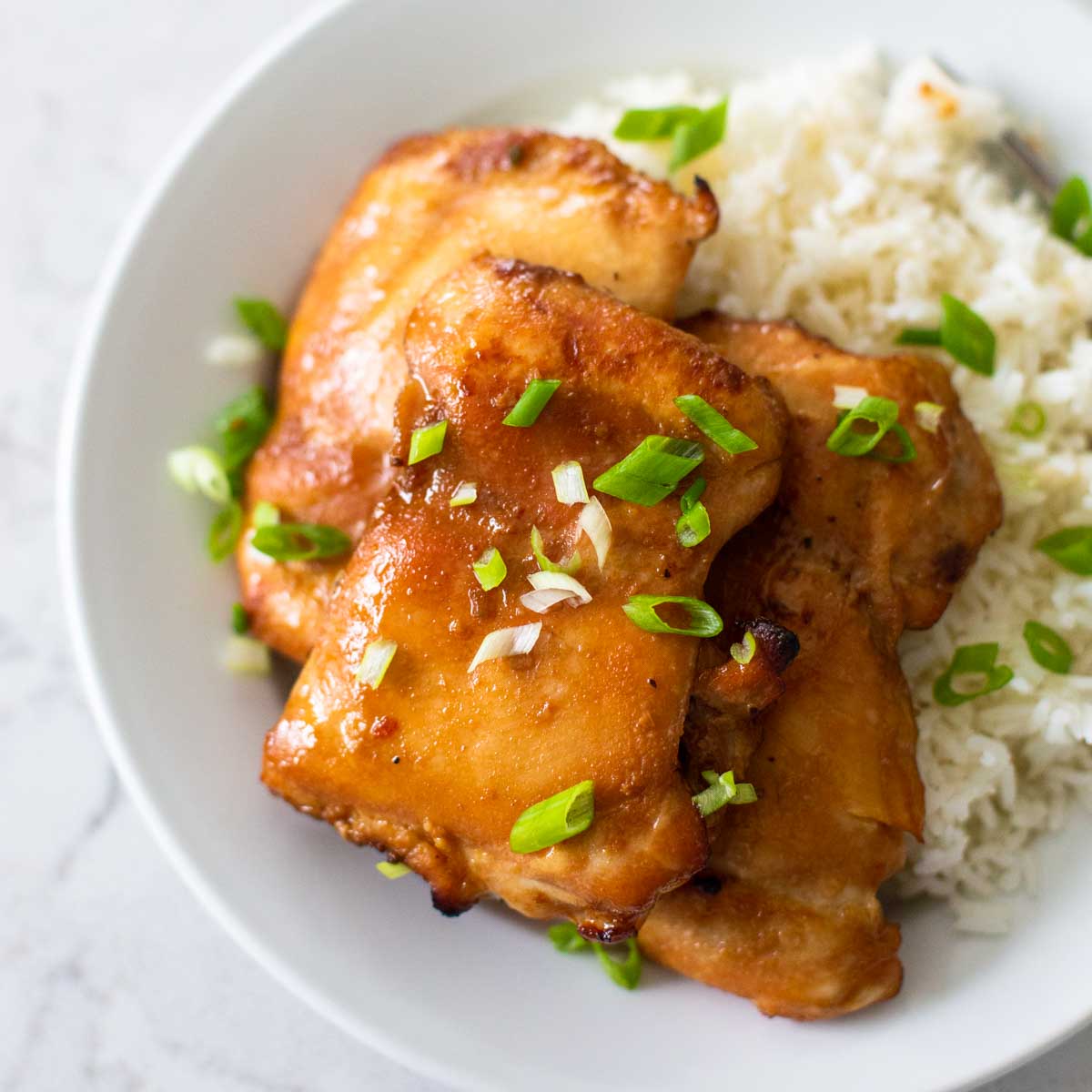 A white plate has jasmine rice and a pile of honey garlic chicken thighs sprinkled with fresh green onions.