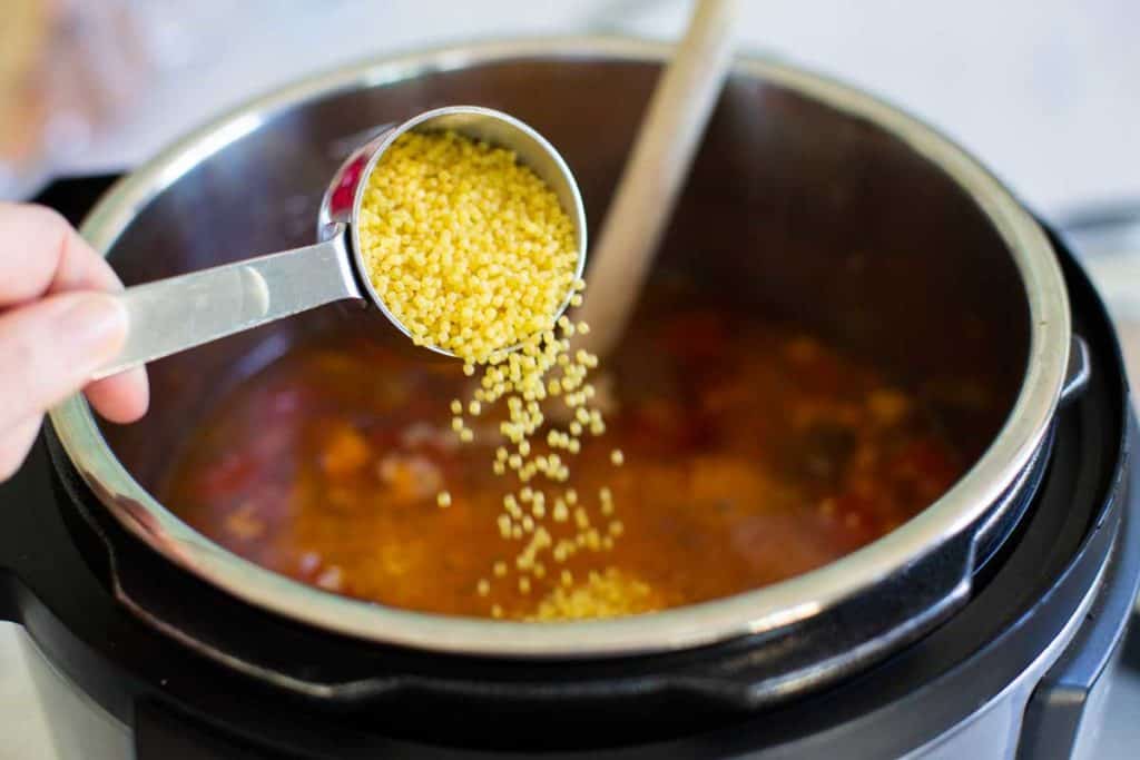 A measuring cup is pouring in the dried pasta into the cooked soup.