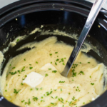A slowcooker with mashed potatoes for a crowd has butter melting on top with fresh chives sprinkled and a serving spoon.