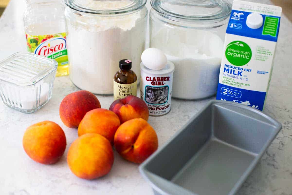 The ingredients for the peach bread are on the kitchen counter. A pile of fresh peaches is in front.