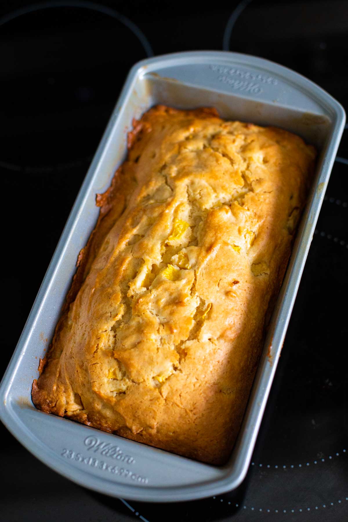 The baked peach bread is in a metal bread pan cooling on the stove top.
