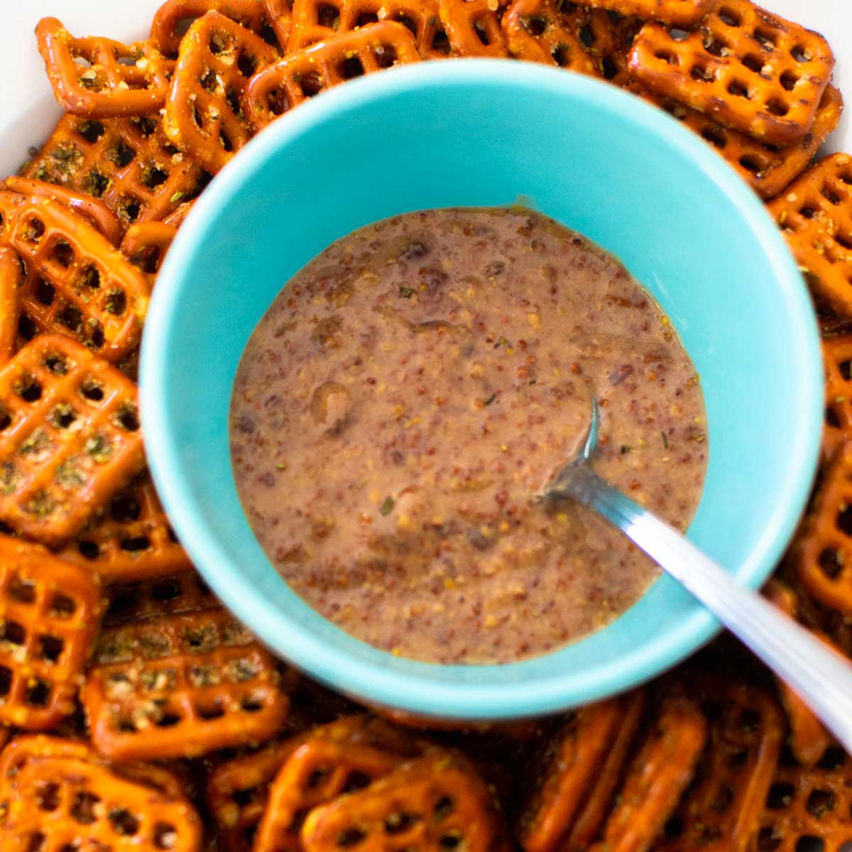A blue bowl filled with raspberry mustard has a spoon and is surrounded by seasoned pretzels.