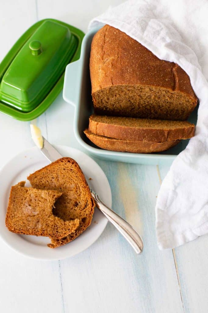 The steakhouse brown bread has been sliced and served in a dish with a towel covering it to keep it warm. A slice has been buttered on a plate and has a bite out of it.