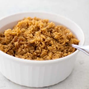 A white serving dish has the beef broth and butter flavored rice with a spoon.