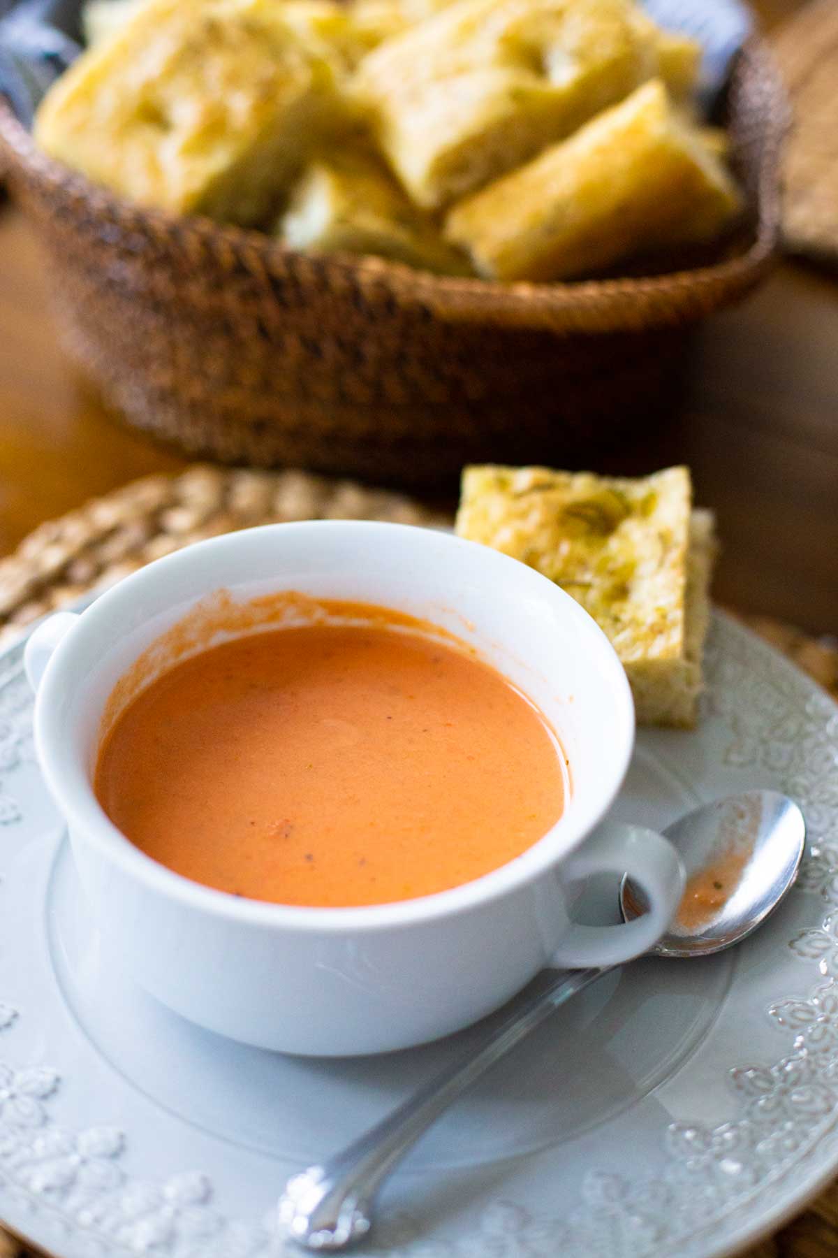 A serving of tomato bisque on a plate next to a basket filled with homemade bread.