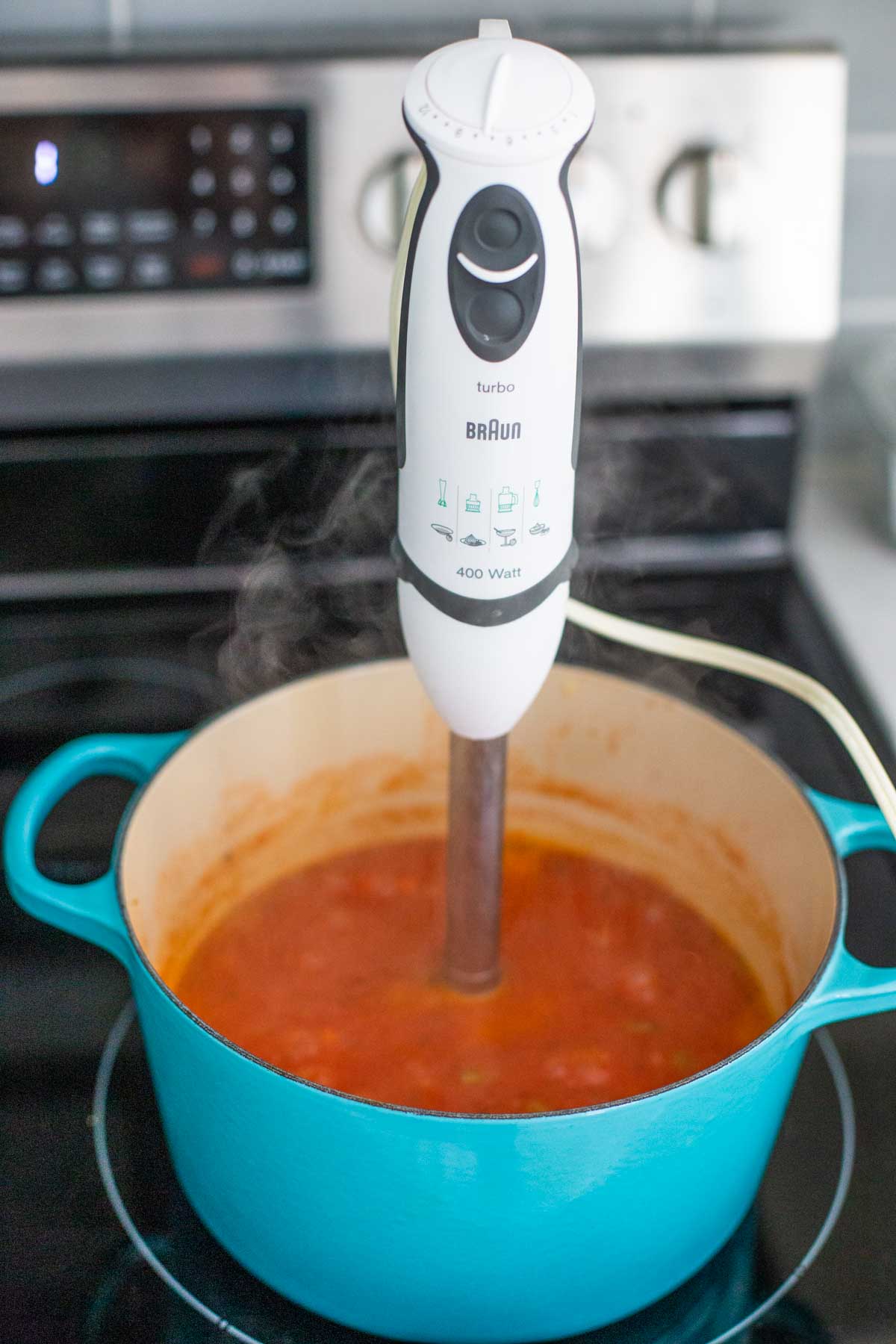 An immersion blender is set inside a blue Le Crueset enameled cast iron soup pot filled with tomato bisque soup.
