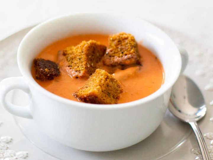 A white soup bowl filled with tomato bisque has brown bread croutons floating on top.