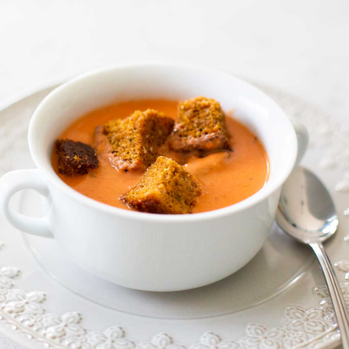 A white soup bowl filled with tomato bisque has brown bread croutons floating on top.