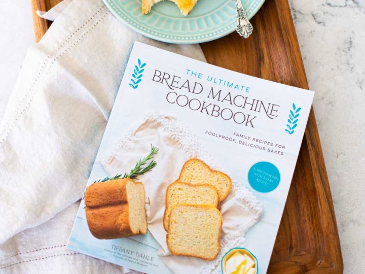 A copy of The Ultimate Bread Machine Cookbook sits on a wooden tray with a napkin and a plate of buttered toast.